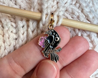 May include: A gold-toned metal charm featuring a black raven perched on a branch with a pink rose. The raven has a small white crescent moon detail on its wing. The charm includes a gold jump ring at the top for attachment to jewelry or accessories.