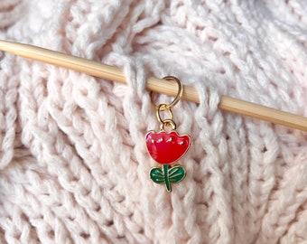 May include: A gold-toned metal stitch marker featuring a red tulip charm with green leaves and stem. The charm is attached to a circular ring, which is positioned on a light-colored wooden knitting needle resting against a soft, pale pink knitted fabric.