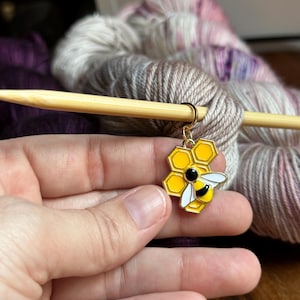 May include: A gold bee-shaped stitch marker with a yellow honeycomb design. The marker is attached to a wooden knitting needle.