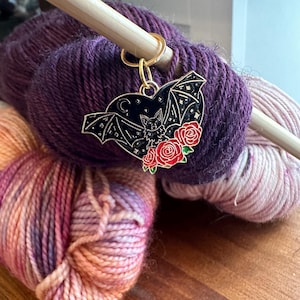 May include: A gold-colored metal bat-shaped stitch marker with a black enamel fill, a bat silhouette, and red roses. The marker is attached to a wooden knitting needle and rests on a skein of purple yarn.