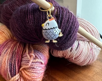 May include: A close-up of colorful yarn skeins with a wooden knitting needle. A gold-colored chicken-shaped stitch marker is attached to the yarn. The yarn is a mix of purple, pink, and orange hues, perfect for knitting or crochet projects.