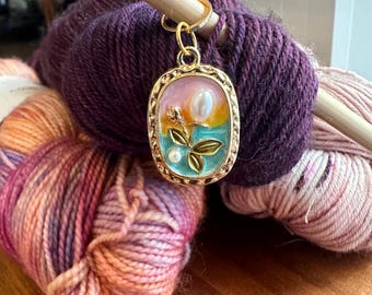 May include: A gold-toned stitch marker with a rectangular, clear resin charm featuring a floral design with pearl accents. The charm has a pink, yellow, and blue background.