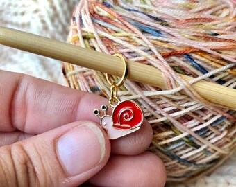 May include: A gold snail stitch marker with a pink body and a red shell. The marker is attached to a wooden knitting needle.