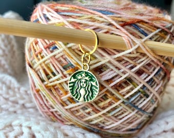 May include: A gold knitting stitch marker with a green Starbucks logo charm. The charm features a mermaid with a crown and the Starbucks logo.