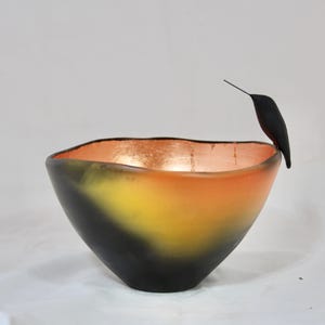 May include: A decorative bowl with a gradient of colors, transitioning from black at the base to yellow and orange, with a copper-colored interior. A black bird figurine perches on the bowl's rim.