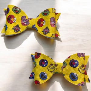 May include: Two yellow hair bows with a repeating pattern of Sesame Street characters. The bows are made of a textured material and feature Elmo, Cookie Monster, and other characters. The bows are a fun accessory.