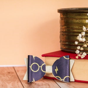 May include: A navy blue and gold bow with a Harry Potter theme, featuring glasses and lightning bolt designs. The bow is set on a stack of books with a brass container and dried flowers in the background. The bow is approximately 4 inches wide.