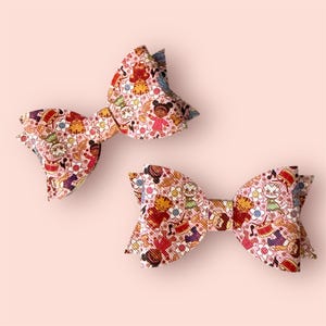 May include: Two pink hair bows with a repeating pattern of cartoon characters, musical instruments, and flowers. The bows are made of a textured material and are set against a light pink background. These bows are perfect for adding a touch of whimsy to any hairstyle.