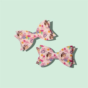 Puede incluir: Dos lazos rosas para el pelo con un estampado repetido de una niña de dibujos animados, bolsas de la compra y caramelos. Los lazos son de un material texturizado sobre un fondo verde claro.