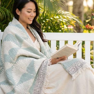 May include: A light blue and white patterned blanket with a floral design and fringe trim. The blanket is draped over a person sitting on a white bench, reading a book. The blanket is made of a soft, textured material.