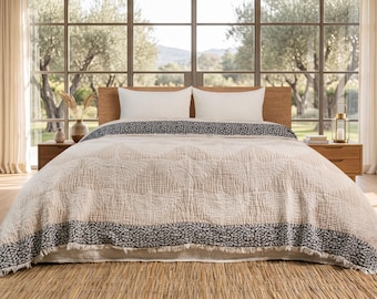May include: A textured beige bedspread with a dark gray floral border and frayed edges, draped over a bed with two white pillows. The bedspread features a subtle quilted pattern and covers the mattress, extending down to the floor.