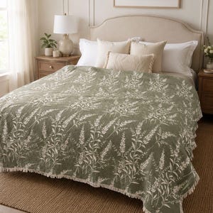 May include: A sage green bedspread featuring a white botanical print of tall grasses and wildflowers. The fabric has a textured, quilted appearance and is finished with a short, knotted fringe along the bottom edge of the coverlet.