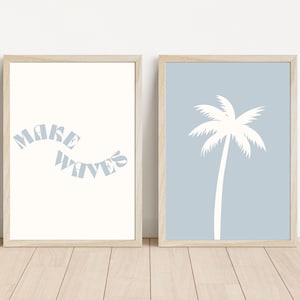 May include: Two framed art prints. One has the text "MAKE WAVES" in light blue on a cream background. The other print features a white palm tree silhouette on a light blue background. Both are in light wood frames.