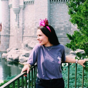 May include: A woman wearing a purple t-shirt and denim shorts stands in front of Cinderella's Castle at Walt Disney World. She is wearing pink mouse ears and smiling. The castle is in the background, and there is a body of water in the foreground.