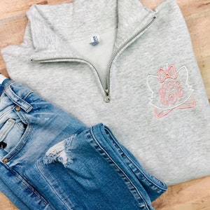 May include: A light gray quarter-zip sweatshirt with a pink and white embroidered cat design and a monogram. The sweatshirt is paired with blue denim jeans with distressed details. The clothing is displayed on a light wood surface.