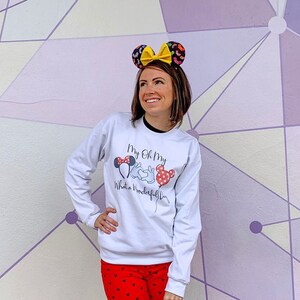May include: A white sweatshirt with a graphic of Mickey Mouse and Minnie Mouse holding hands with the text "My Oh My, What a Wonderful Day!" printed on the front. The person is wearing red pants with a white polka dot pattern and a pair of black and yellow mouse ears.