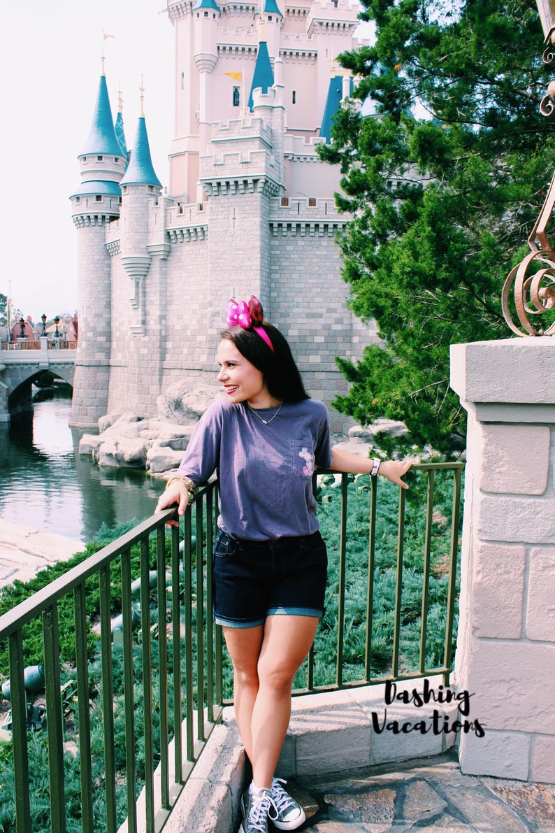 May include: A woman wearing a purple t-shirt and denim shorts stands in front of Cinderella's Castle at Walt Disney World. She is wearing pink mouse ears and smiling. The castle is in the background, and there is a body of water in the foreground.