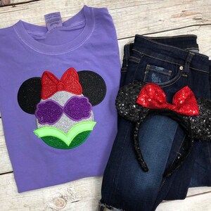 May include: Purple t-shirt with a glittery, green, purple, and red embroidered design of a Disney character with a bow. The design is in the shape of Mickey Mouse ears. A pair of black and red sequined Mickey Mouse ears are on a pair of blue jeans.