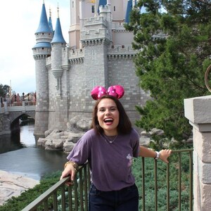 May include: A woman wearing a purple t-shirt and denim shorts smiles in front of Cinderella's Castle at Walt Disney World. She is wearing pink Minnie Mouse ears with white polka dots.
