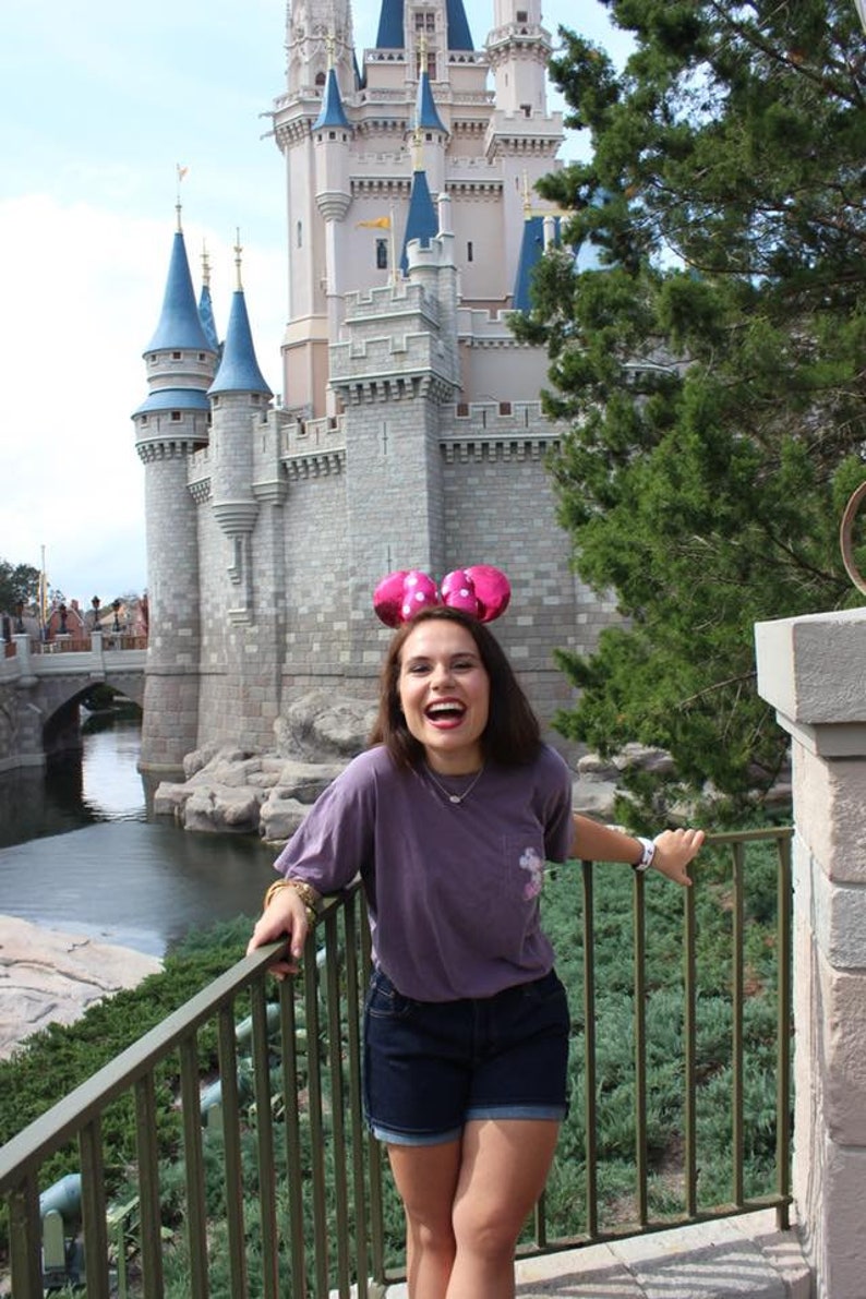 May include: A woman wearing a purple t-shirt and denim shorts smiles in front of Cinderella's Castle at Walt Disney World. She is wearing pink Minnie Mouse ears with white polka dots.
