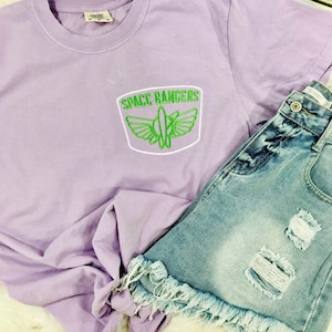 May include: A lavender t-shirt with a green embroidered patch that says "Space Rangers" with a rocket ship and wings. The t-shirt is paired with a light blue denim skirt with frayed edges and distressed details.