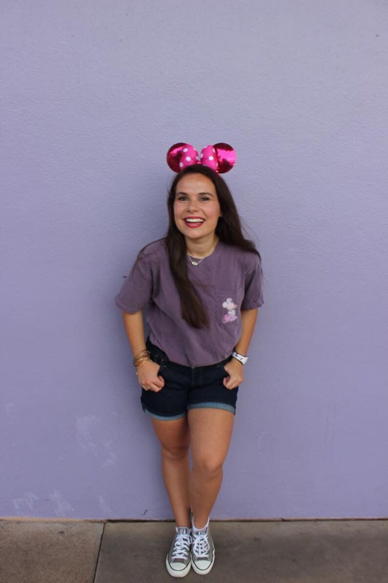 May include: A woman wearing a purple t-shirt with a white Mickey Mouse graphic, denim shorts, and white sneakers. She is smiling and wearing pink Minnie Mouse ears with white polka dots.