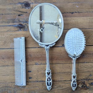 May include: A vintage vanity set comprising a round hand mirror, a hairbrush, and a comb. The set has ornate silver-coloured handles with heart and floral designs. The comb and brush have white bristles. The items are arranged on a wooden surface.