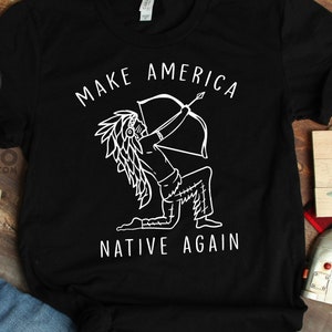 May include: Black t-shirt with the text "MAKE AMERICA NATIVE AGAIN" and a white graphic of a Native American figure with a bow and arrow. The shirt is paired with blue jeans on a wooden surface.