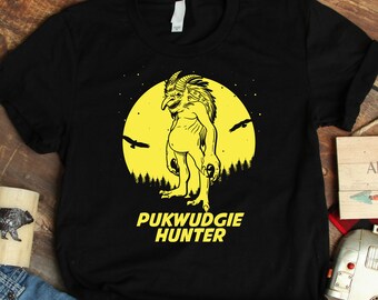 Può includere: Maglietta nera con una grafica gialla di una creatura con una testa grande e una luna piena. Il testo "PUKWUDGIE HUNTER" è sotto la grafica. La maglietta è su una superficie di legno con un paio di jeans blu, una tazza di legno, un piccolo camper giocattolo e un cartello di legno che dice "NOT ALL WHO WANDER ARE LOST".