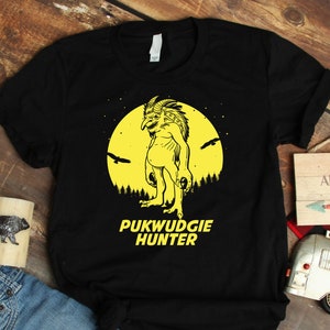 May include: Black t-shirt with a yellow graphic of a creature with a large head and a full moon. The text "PUKWUDGIE HUNTER" is below the graphic. The t-shirt is on a wooden surface with a pair of blue jeans, a wooden mug, a small toy camper, and a wooden sign that says "NOT ALL WHO WANDER ARE LOST".