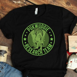 May include: Black t-shirt with a green circle graphic that says "Pukwudgie Research Team". The graphic features a green creature with a large head and small body. The t-shirt is on a wooden surface with a small wooden sign that says "Not all who wander are lost".