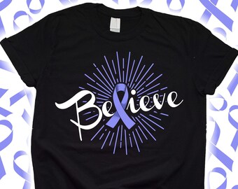 Puede incluir: Camiseta negra con la palabra "Believe" en escritura blanca, una cinta morada clara y líneas radiantes. El diseño está centrado en la camiseta. Cintas moradas claras en el fondo.