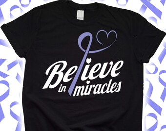 Puede incluir: Camiseta negra con la frase "Believe in miracles" en escritura blanca, acentuada por una cinta y un corazón de color lavanda. La camiseta es de cuello redondo.