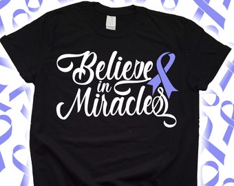 Puede incluir: Camiseta negra con la frase "Believe in Miracles" en blanco y un lazo morado claro. La camiseta tiene mangas cortas y cuello redondo. Cintas moradas claras en el fondo.