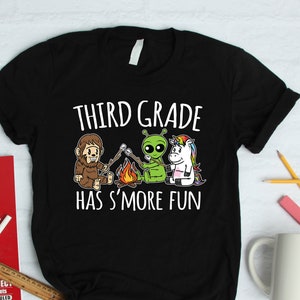 May include: Black t-shirt with the text "THIRD GRADE HAS S'MORE FUN." The graphic features a cartoon Bigfoot, an alien, and a unicorn roasting marshmallows over a campfire. A fun design for third grade students.