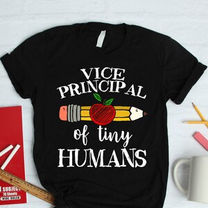 May include: Black t-shirt with the text "VICE PRINCIPAL of tiny HUMANS" in white. The design includes a pencil with an apple. School supplies like a notebook, ruler, and pencils are next to the shirt.