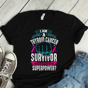 Puede incluir: Camiseta negra con un diseño de cinta turquesa y rosa y el texto "Soy un superviviente de cáncer de tiroides ¿Cuál es tu superpoder?"