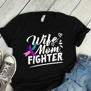 Puede incluir: Camiseta negra con las palabras "Wife, Mom, Fighter" en letras blancas. La camiseta presenta un diseño de cinta turquesa y morada. Zapatillas grises y vaqueros azules junto a la camiseta.