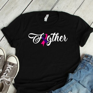 Puede incluir: Camiseta negra con la palabra "Fighter" en escritura blanca, con un diseño de cinta turquesa, morada y rosa. La camiseta se combina con zapatillas grises y vaqueros azules, creando un look casual.