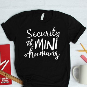 以下が含まれることがあります： 白い文字で「Security of MINI humans」と書かれた黒いTシャツ。赤いノート、チョーク、定規など、学用品と一緒に白い面にシャツが表示されています。