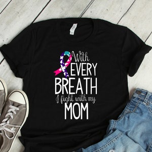 Puede incluir: Camiseta negra con el texto "With Every Breath I fight with my MOM" y un lazo rosa, azul y blanco. La camiseta se combina con zapatillas grises y vaqueros azules.