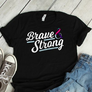 Puede incluir: Camiseta negra con el texto "Brave & Strong" en blanco con un símbolo de cinta azul y morada. La camiseta está colocada sobre un fondo de madera blanca con un par de zapatillas grises y unos vaqueros azules.