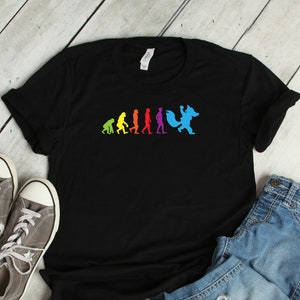 Può includere: Maglietta nera con una grafica dell'evoluzione dell'uomo che termina con una volpe blu. La grafica è in colori arcobaleno.