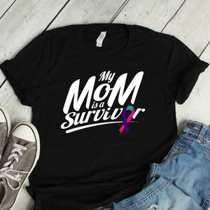 Puede incluir: Camiseta negra con texto blanco que dice "My Mom is a Survivor" y una cinta morada, turquesa y rosa.