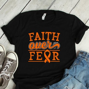 May include: Black t-shirt with the words "FAITH over FEAR" in orange lettering and an orange ribbon graphic. The shirt is paired with gray sneakers and blue jeans on a light wood background.