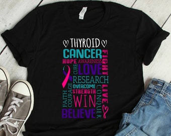 Può includere: Maglietta nera con un nastro rosa e le parole "Cancro alla tiroide" in una colonna verticale. Le parole "Speranza, Consapevolezza, Cura, Amore, Ricerca, Superare, Coraggio, Forza, Vincere, Conquistare, Fede, Vivere, Credere" sono disposte attorno al nastro e alle parole "Cancro alla tiroide".