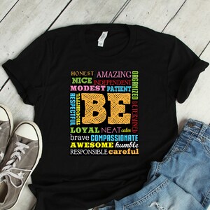 May include: Black t-shirt with the word "BE" in orange and yellow, surrounded by positive adjectives in various colors. Gray sneakers and blue jeans are next to the shirt on a light wood surface.