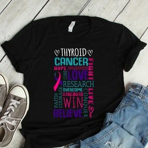 Puede incluir: Camiseta negra con una cinta rosa y las palabras "Cáncer de tiroides" en una columna vertical. Las palabras "Esperanza, Conciencia, Cura, Amor, Investigación, Superar, Coraje, Fuerza, Ganar, Conquistar, Fe, Vivir, Creer" están dispuestas alrededor de la cinta y las palabras "Cáncer de tiroides".