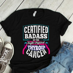 Puede incluir: Camiseta negra con el texto "CERTIFIED BADASS in the Fight Against Thyroid Cancer" en letras blancas y turquesas. El diseño incluye una cinta rosa y morada. Zapatillas grises y vaqueros azules junto a la camiseta.