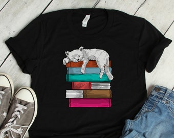 May include: Black t-shirt with a white cat sleeping on a stack of colorful books. The cat is facing left and has its paws hanging off the top book. The books are stacked on top of each other and have different colors and designs.